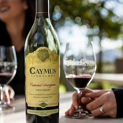 Caymus Vineyards wine bottle with a glass of red wine on a blurred outdoor background